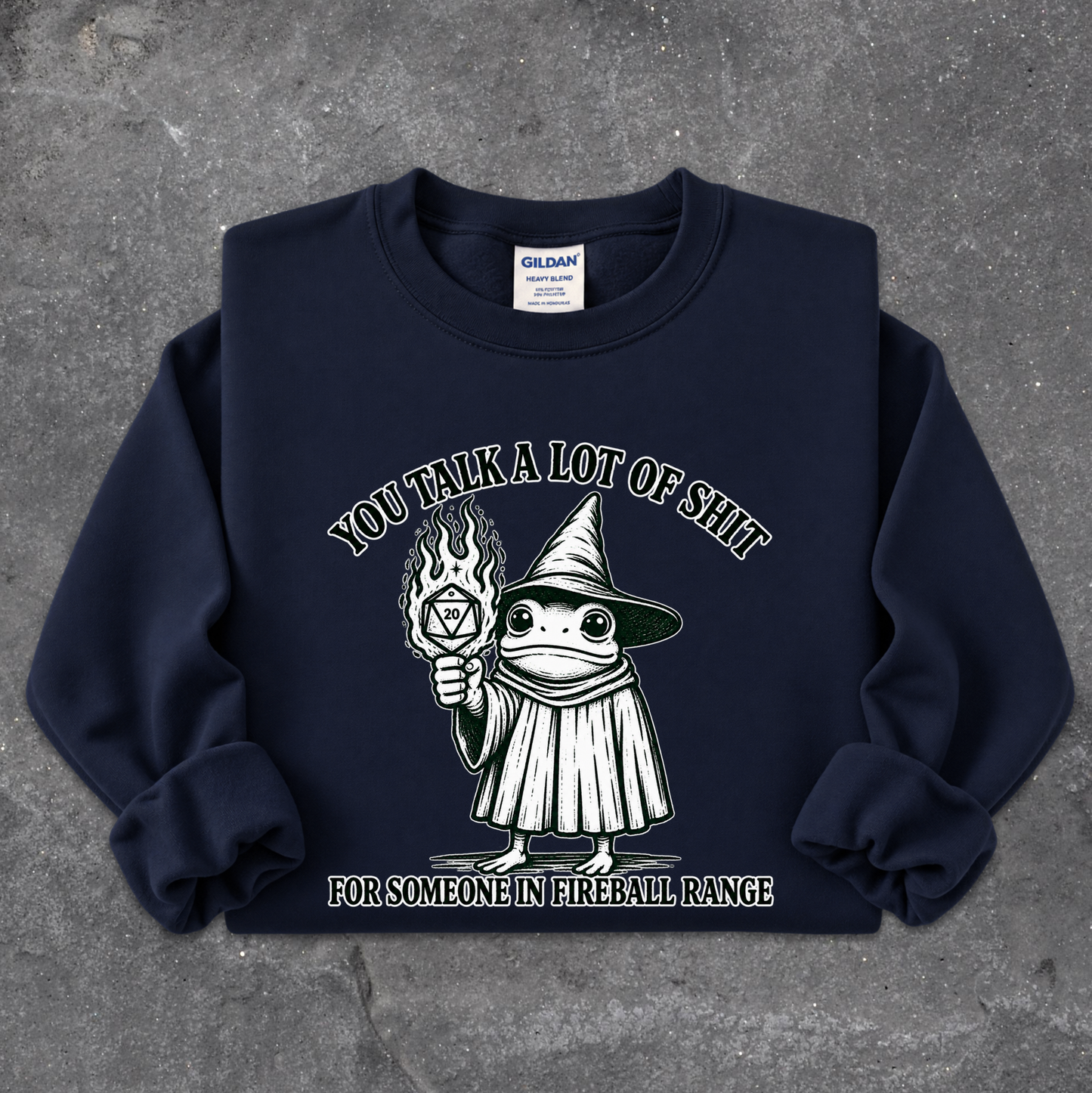A navy blue crewneck sweatshirt with a graphic of a cartoon frog in a wizard hat holding a flaming torch, with the text 'YOU TALK A LOT OF SHIT FOR SOMEONE IN FIREBALL RANGE'. The sweatshirt is laid flat on a textured gray concrete surface.