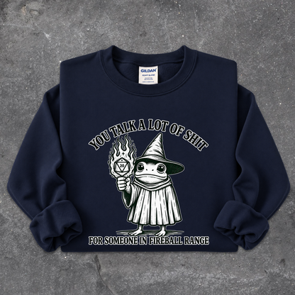 A navy blue crewneck sweatshirt with a graphic of a cartoon frog in a wizard hat holding a flaming torch, with the text 'YOU TALK A LOT OF SHIT FOR SOMEONE IN FIREBALL RANGE'. The sweatshirt is laid flat on a textured gray concrete surface.