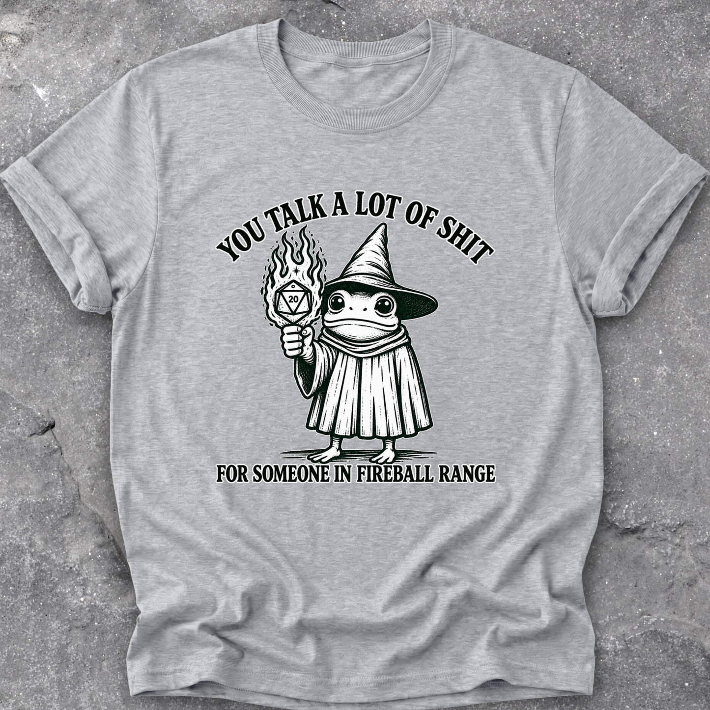 A heather gray short-sleeve t-shirt with a black graphic of a frog in a wizard hat holding a flaming orb, with the text 'YOU TALK A LOT OF SHIT FOR SOMEONE IN FIREBALL RANGE', laid flat on a textured gray surface.