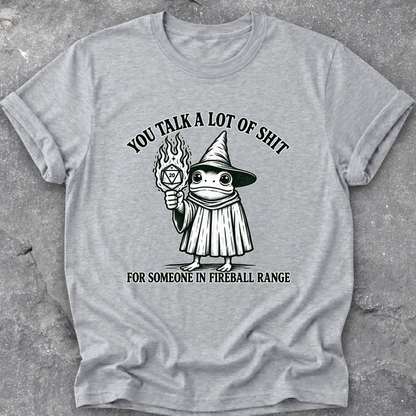 A heather gray short-sleeve t-shirt with a black graphic of a frog in a wizard hat holding a flaming orb, with the text 'YOU TALK A LOT OF SHIT FOR SOMEONE IN FIREBALL RANGE', laid flat on a textured gray surface.