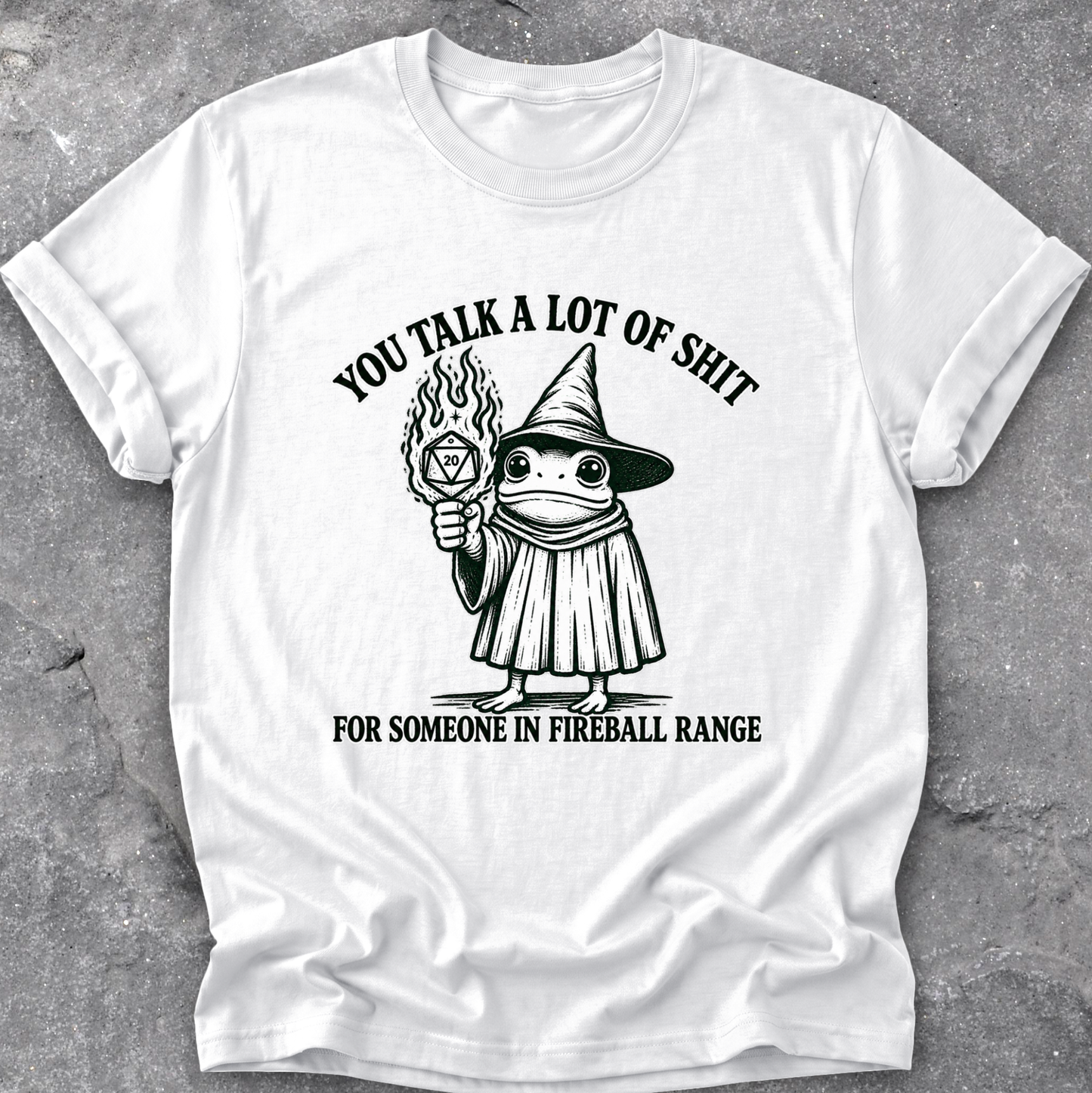 A white short-sleeve crewneck t-shirt with a black graphic print of a cartoon frog in a wizard hat holding a flaming orb, with the text 'YOU TALK A LOT OF SHIT' above and 'FOR SOMEONE IN FIREBALL RANGE' below, laid flat on a gray textured surface.
