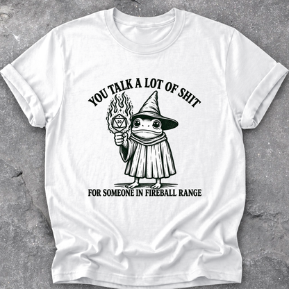 A white short-sleeve crewneck t-shirt with a black graphic print of a cartoon frog in a wizard hat holding a flaming orb, with the text 'YOU TALK A LOT OF SHIT' above and 'FOR SOMEONE IN FIREBALL RANGE' below, laid flat on a gray textured surface.