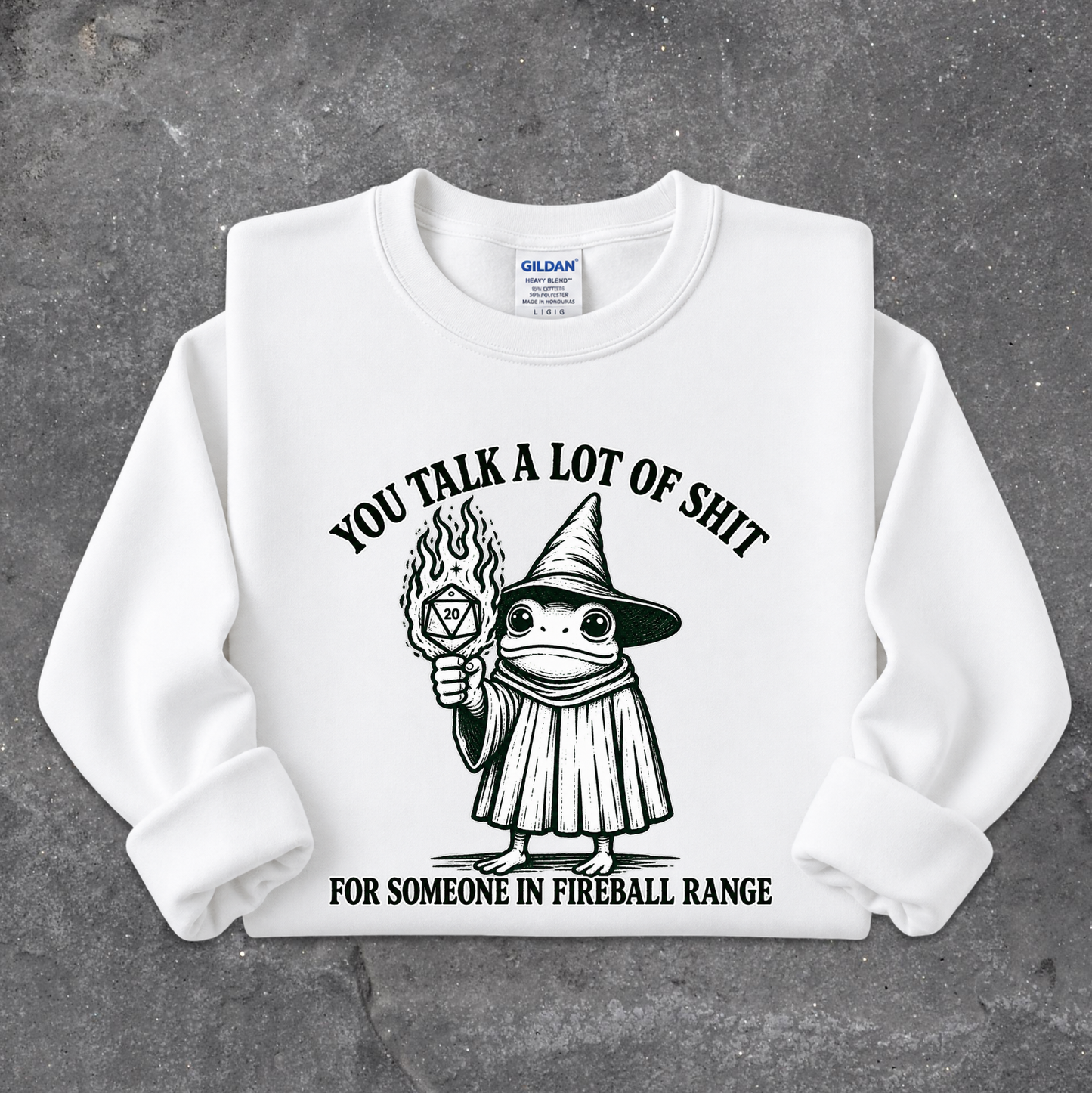 A white crewneck sweatshirt with a black graphic of a cartoon frog in a wizard hat holding a flaming torch, with the text 'YOU TALK A LOT OF SHIT FOR SOMEONE IN FIREBALL RANGE'. The sweatshirt is laid flat on a textured gray background.