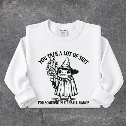 A white crewneck sweatshirt with a black graphic of a cartoon frog in a wizard hat holding a flaming torch, with the text 'YOU TALK A LOT OF SHIT FOR SOMEONE IN FIREBALL RANGE'. The sweatshirt is laid flat on a textured gray background.