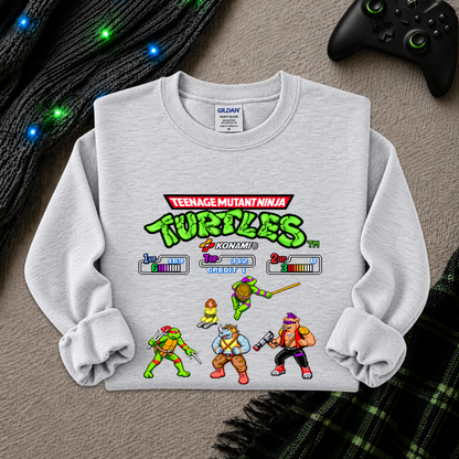 A light gray heather crewneck sweatshirt featuring a colorful Teenage Mutant Ninja Turtles video game-style graphic on the front, laid flat on a textured gray surface with a black controller, knit blanket, and plaid fabric nearby.
