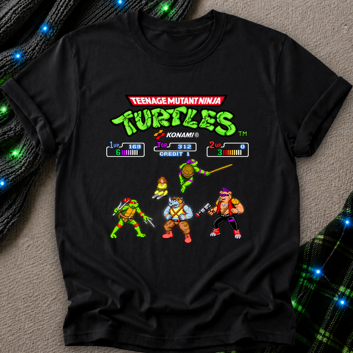 A black short-sleeve graphic T-shirt laid flat on a textured gray surface with decorative blue lights and plaid fabric accents. The shirt features a colorful Teenage Mutant Ninja Turtles video game-style print with the franchise title, score HUD elements, and four turtle characters.