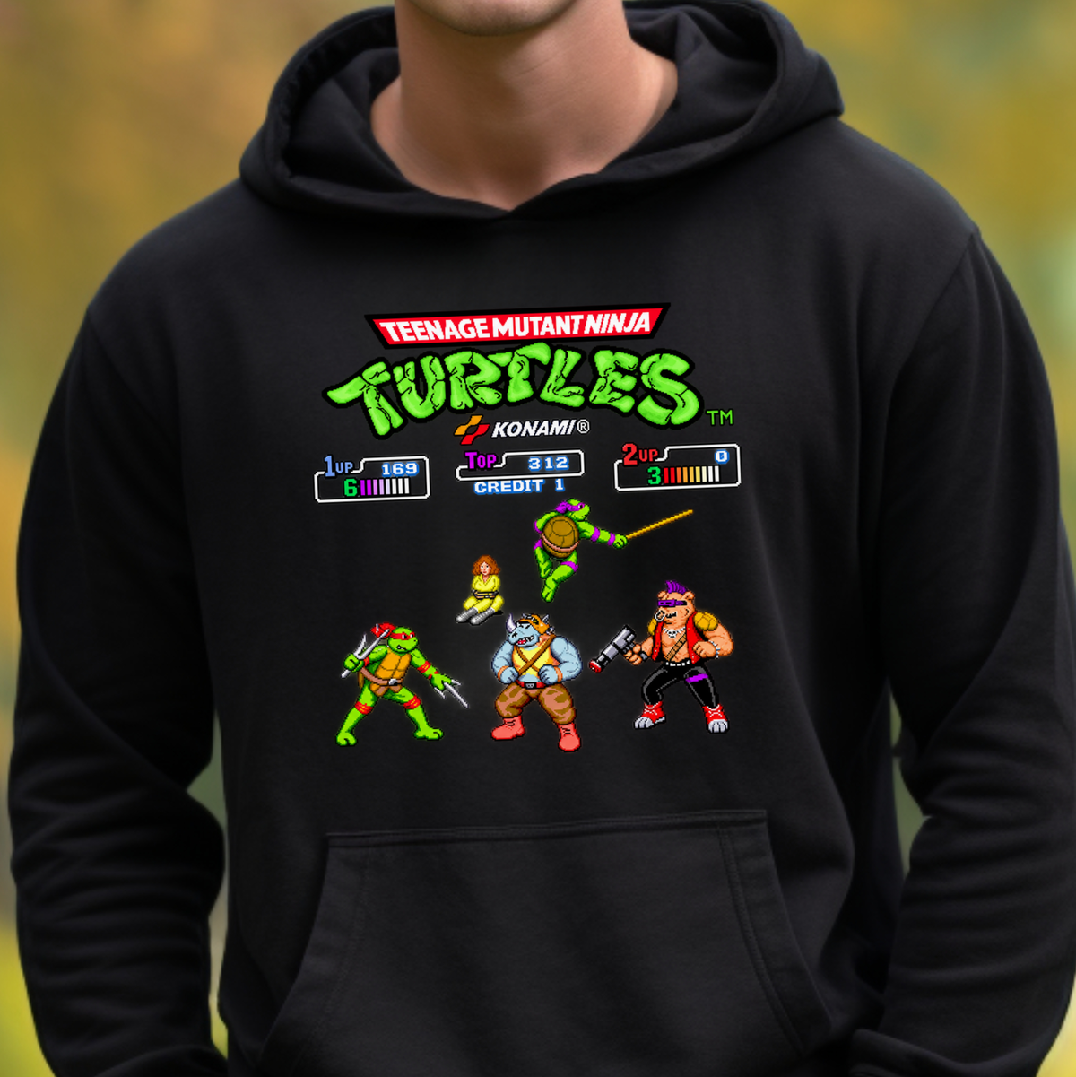 A person wearing a black hoodie with a colorful Teenage Mutant Ninja Turtles video game-style graphic on the chest, including the Konami logo and character illustrations, against a blurred outdoor background.