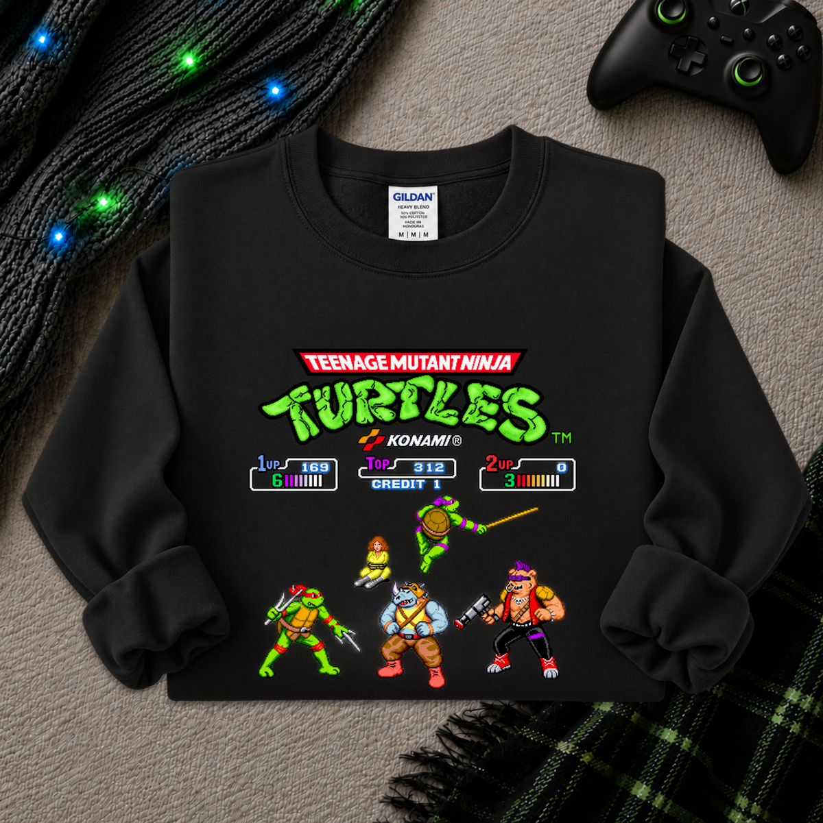 A black long-sleeve crewneck sweatshirt featuring a colorful Teenage Mutant Ninja Turtles video game screen graphic on the front, laid flat on a gray textured surface with a black controller, knit cable, and plaid blanket nearby.