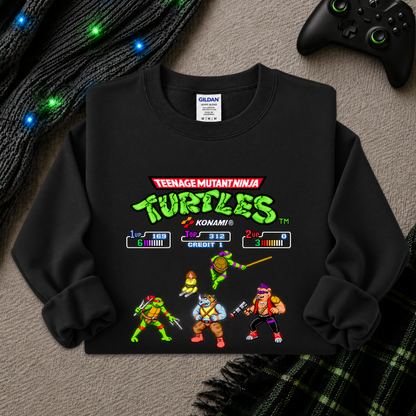 A black long-sleeve crewneck sweatshirt featuring a colorful Teenage Mutant Ninja Turtles video game screen graphic on the front, laid flat on a gray textured surface with a black controller, knit cable, and plaid blanket nearby.