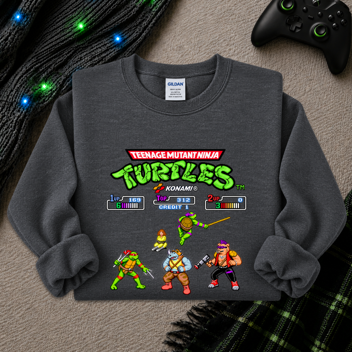 A dark gray crewneck sweatshirt featuring a retro-style Teenage Mutant Ninja Turtles video game screen graphic on the front, styled in a cozy flat-lay with a black controller, glowing holiday lights, and plaid fabric in the background.