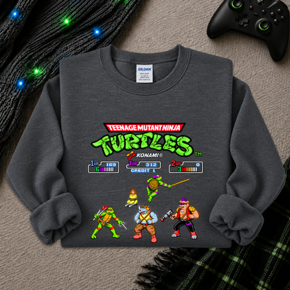 A dark gray crewneck sweatshirt featuring a retro-style Teenage Mutant Ninja Turtles video game screen graphic on the front, styled in a cozy flat-lay with a black controller, glowing holiday lights, and plaid fabric in the background.
