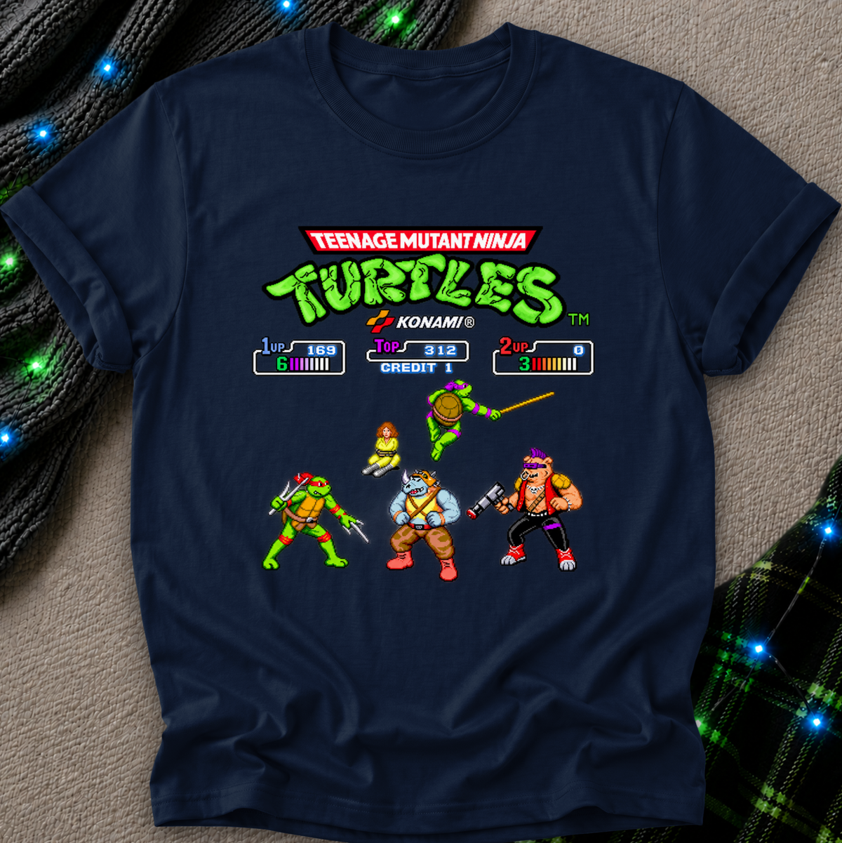 A navy blue short-sleeve t-shirt featuring a colorful retro video game screen graphic of the Teenage Mutant Ninja Turtles on the chest, laid flat on a textured gray surface with decorative green lights and plaid fabric in the corners.