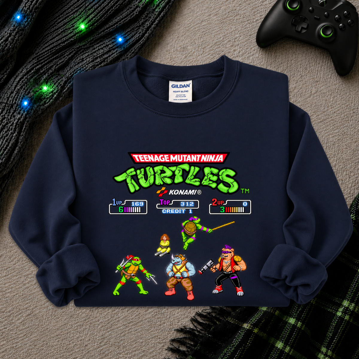 A navy blue crewneck sweatshirt featuring a retro video game screen graphic for Teenage Mutant Ninja Turtles, laid flat on a gray textured surface with a black game controller and glowing holiday lights in the background.