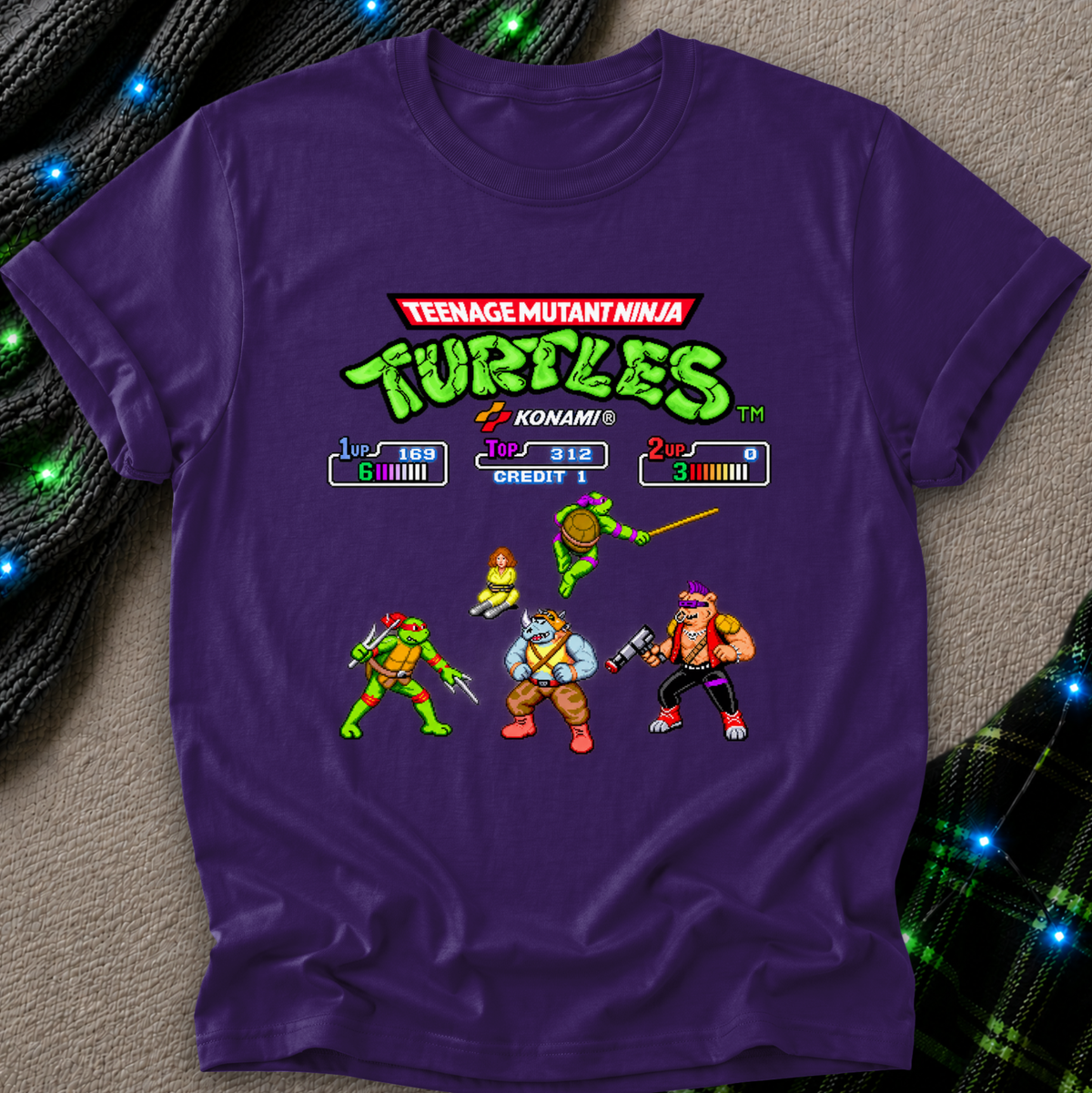 A purple short-sleeve t-shirt featuring a retro video game-style graphic of the Teenage Mutant Ninja Turtles with Konami game-screen elements and character sprites, laid flat on a textured beige surface with decorative blue lights.
