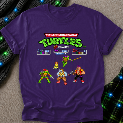 A purple short-sleeve t-shirt featuring a retro video game-style graphic of the Teenage Mutant Ninja Turtles with Konami game-screen elements and character sprites, laid flat on a textured beige surface with decorative blue lights.