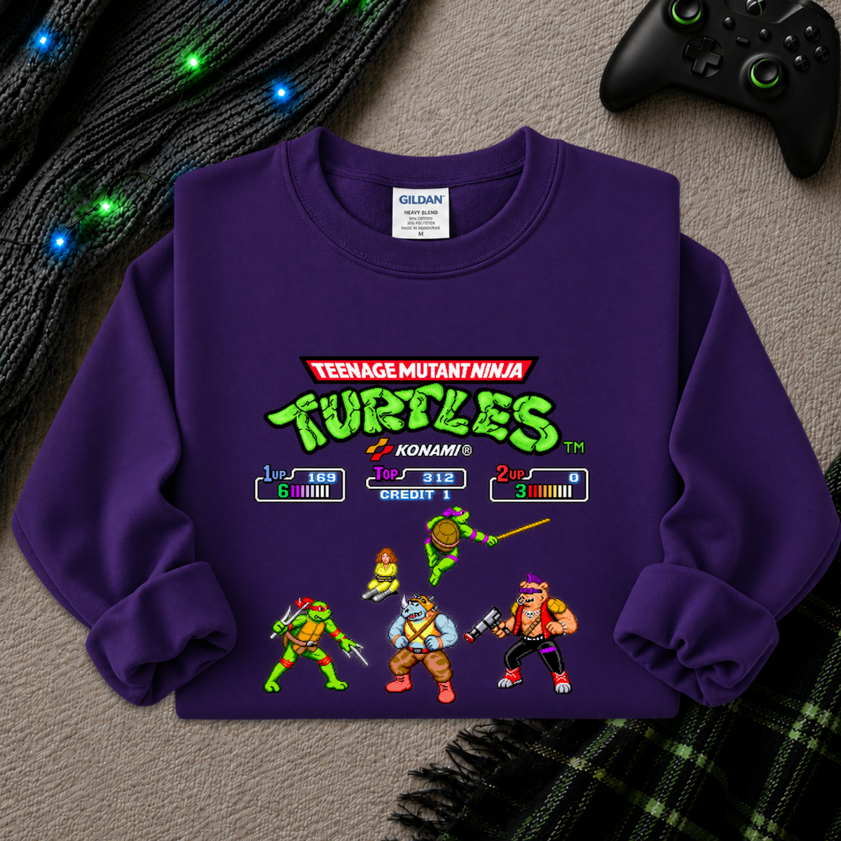 A purple long-sleeve crewneck sweatshirt featuring a retro video game-style Teenage Mutant Ninja Turtles graphic, laid flat on a gray textured surface with a black controller, knit sweater, and plaid blanket nearby.
