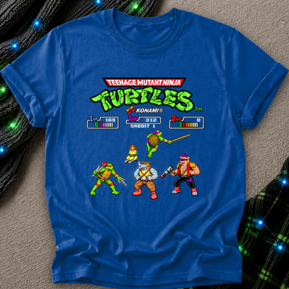 A royal blue short-sleeve t-shirt featuring a retro video game-style graphic of the Teenage Mutant Ninja Turtles and their friends from Konami, laid flat on a textured gray surface with decorative bokeh lights.