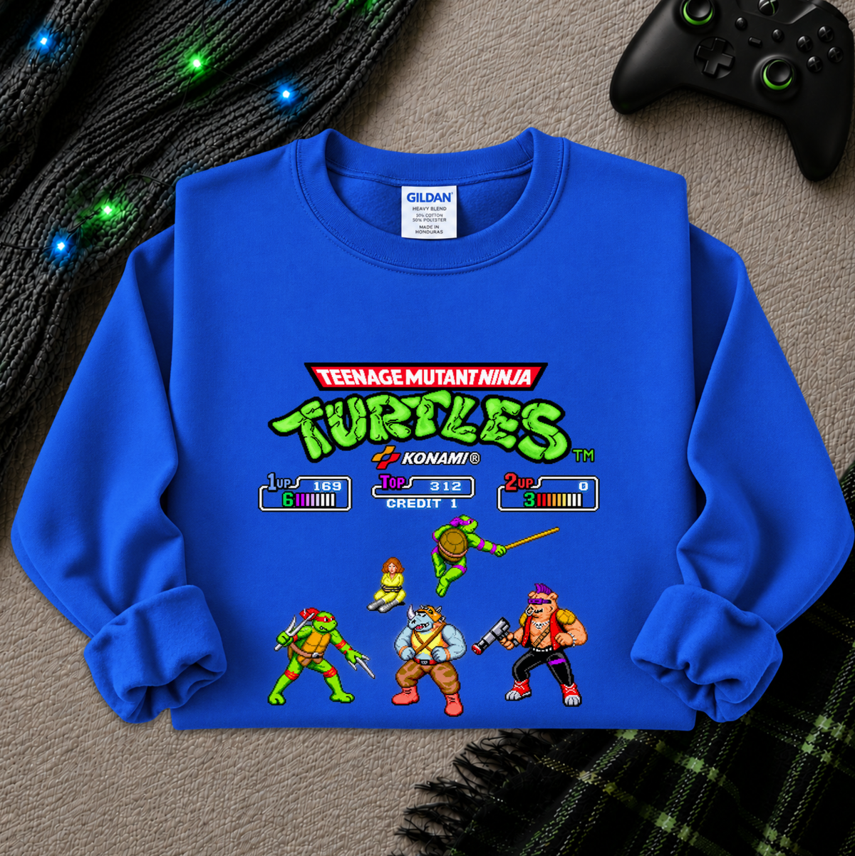 A royal blue Gildan crewneck sweatshirt featuring a retro 8-bit video game screen graphic of the Teenage Mutant Ninja Turtles, laid flat on a gray textured surface with a black controller and plaid blanket as props.