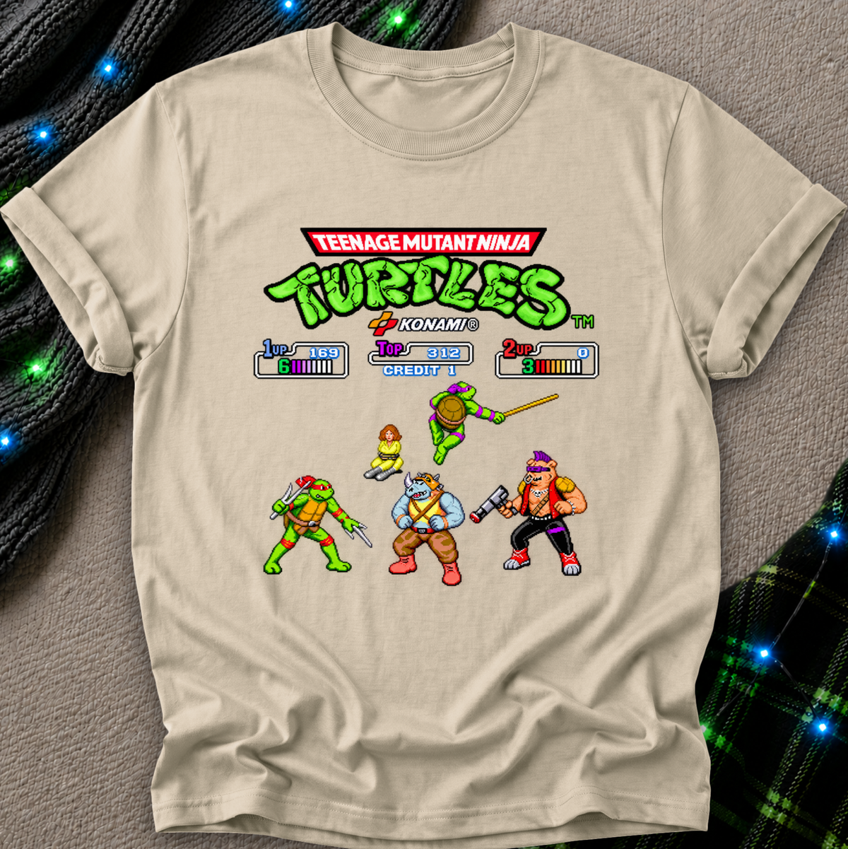 A beige graphic t-shirt featuring a retro 8-bit video game screen design from the Teenage Mutant Ninja Turtles Komatsu 3D. The shirt is laid flat on a gray textured surface with decorative black ribbed fabric and blue fairy lights in the corners.