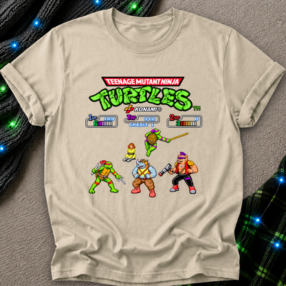 A beige graphic t-shirt featuring a retro 8-bit video game screen design from the Teenage Mutant Ninja Turtles Komatsu 3D. The shirt is laid flat on a gray textured surface with decorative black ribbed fabric and blue fairy lights in the corners.