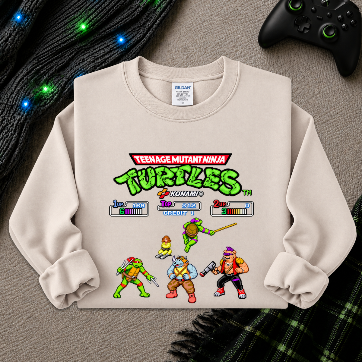 A cream-colored crewneck sweatshirt featuring a colorful retro-style Teenage Mutant Ninja Turtles video game screen graphic, laid flat on a gray textured surface with a black controller, green cable, and plaid blanket as props.