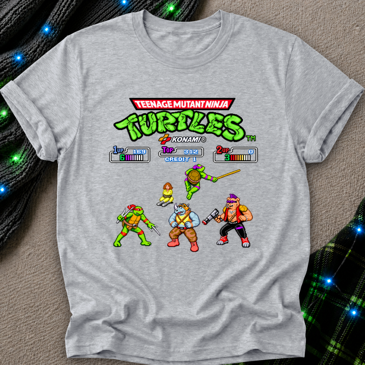 A light heather gray short-sleeve t-shirt featuring a colorful retro video game-style graphic of the Teenage Mutant Ninja Turtles. The shirt is laid flat on a textured beige surface with decorative black cords and blue fairy lights in the background.