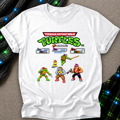 A white short-sleeve crewneck t-shirt featuring a colorful retro Teenage Mutant Ninja Turtles video game graphic on the front, laid flat on a textured gray surface with decorative black cords and blue fairy lights in the background.