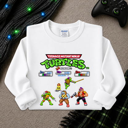 A white long-sleeve crewneck sweatshirt featuring a colorful Teenage Mutant Ninja Turtles video game-style graphic on the front, laid flat on a gray textured surface with a black controller, black cable, and green plaid fabric nearby.