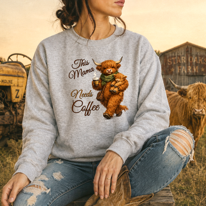 woman wearing a gray sweatshirt with a highland cow holding her calf and a cup of coffee that says this mama needs coffee