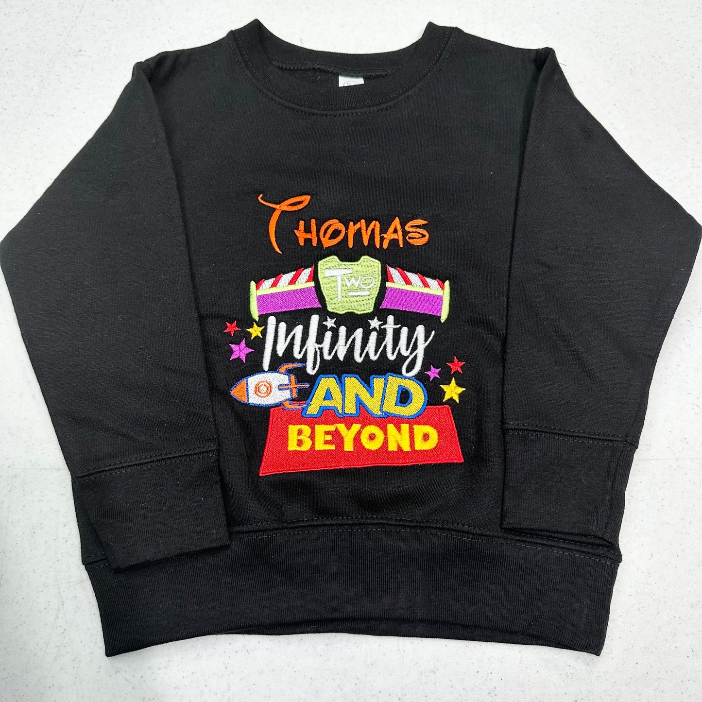 A black long-sleeve crewneck sweatshirt featuring a colorful graphic print on the front with the text 'Thomas Infinity AND BEYOND' and space-themed illustrations, laid flat on a light gray surface.