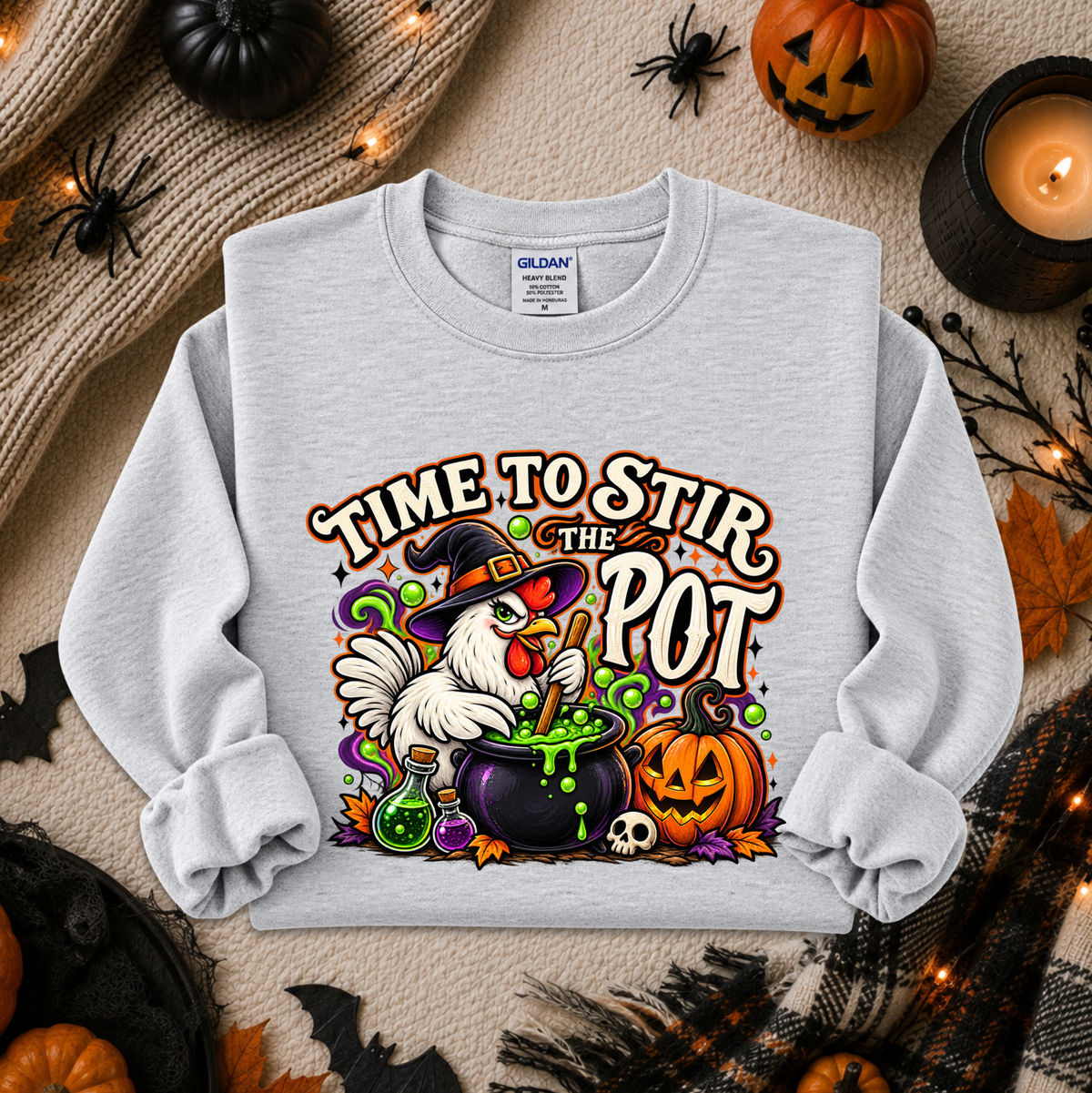 A light gray long-sleeve sweatshirt with a colorful Halloween graphic reading 'TIME TO STIR THE POT,' featuring a white chicken in a witch hat beside a cauldron, pumpkin, skull, and potion bottles. The sweatshirt is laid flat on a cozy fall-themed background with pumpkins, spider decorations, a candle, and autumn leaves.