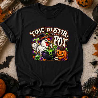 A black short-sleeve graphic T-shirt laid flat on a cozy Halloween-themed background. The shirt features a colorful illustration of a white chicken in a witch hat stirring a cauldron of green potion, with pumpkins, a skull, and the text 'TIME TO STIR THE POT'.