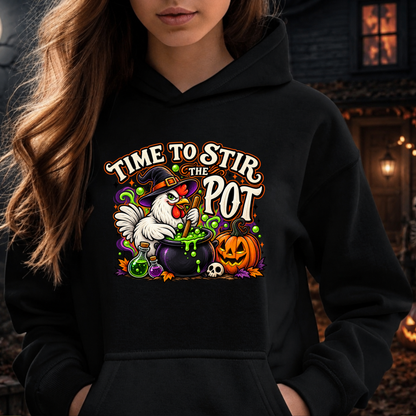 A woman wearing a black hoodie with a colorful Halloween graphic that reads 'TIME TO STIR THE POT' around an illustration of a white chicken in a witch hat stirring a cauldron with green slime, next to a jack-o'-lantern pumpkin.