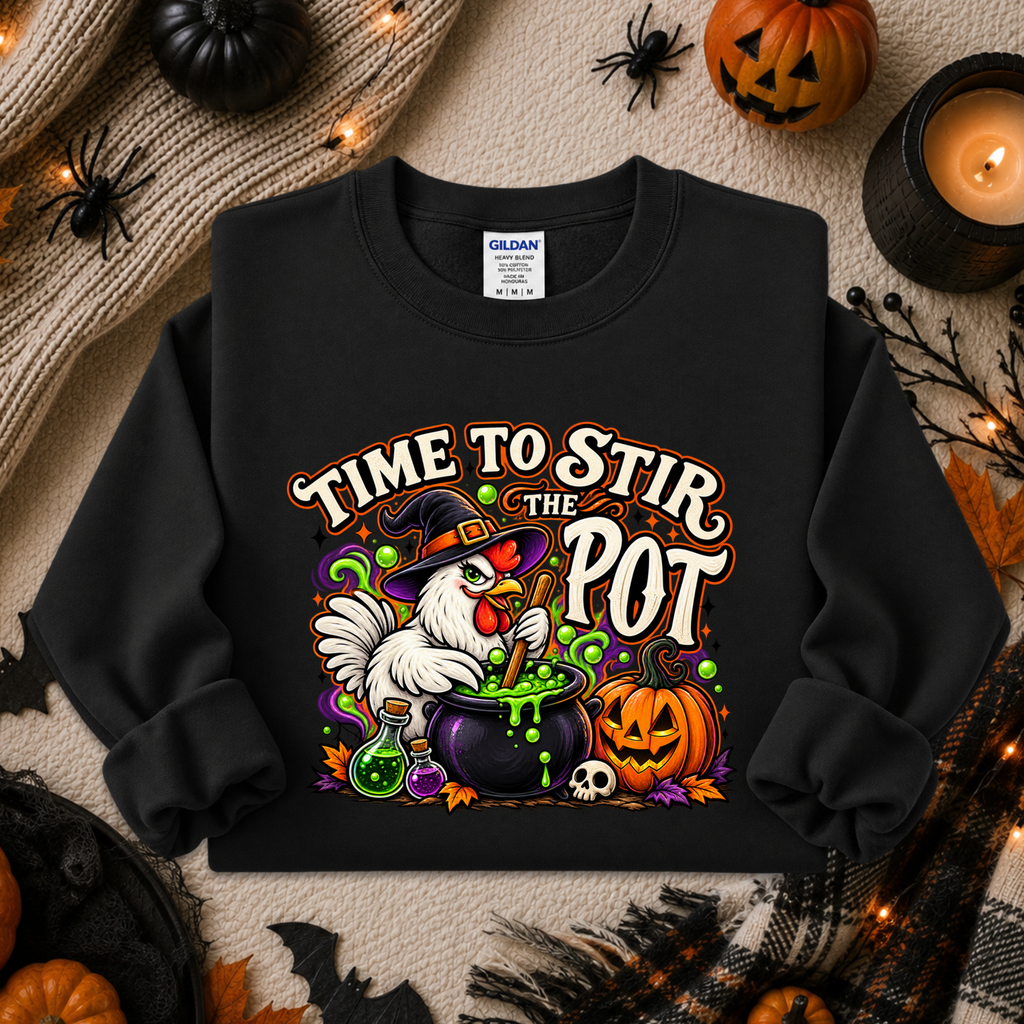 A black long-sleeve crewneck sweatshirt with a colorful Halloween graphic of a white chicken stirring a cauldron of green potion, surrounded by pumpkins, a skull, and the text 'TIME TO STIR THE POT'. The sweatshirt is displayed in a flat-lay arrangement with autumn decorations including a candle, spiderwebs, and pumpkins on a beige knit blanket.