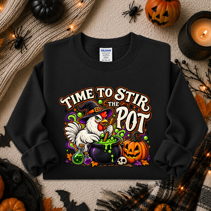 A black long-sleeve crewneck sweatshirt with a colorful Halloween graphic of a white chicken stirring a cauldron of green potion, surrounded by pumpkins, a skull, and the text 'TIME TO STIR THE POT'. The sweatshirt is displayed in a flat-lay arrangement with autumn decorations including a candle, spiderwebs, and pumpkins on a beige knit blanket.