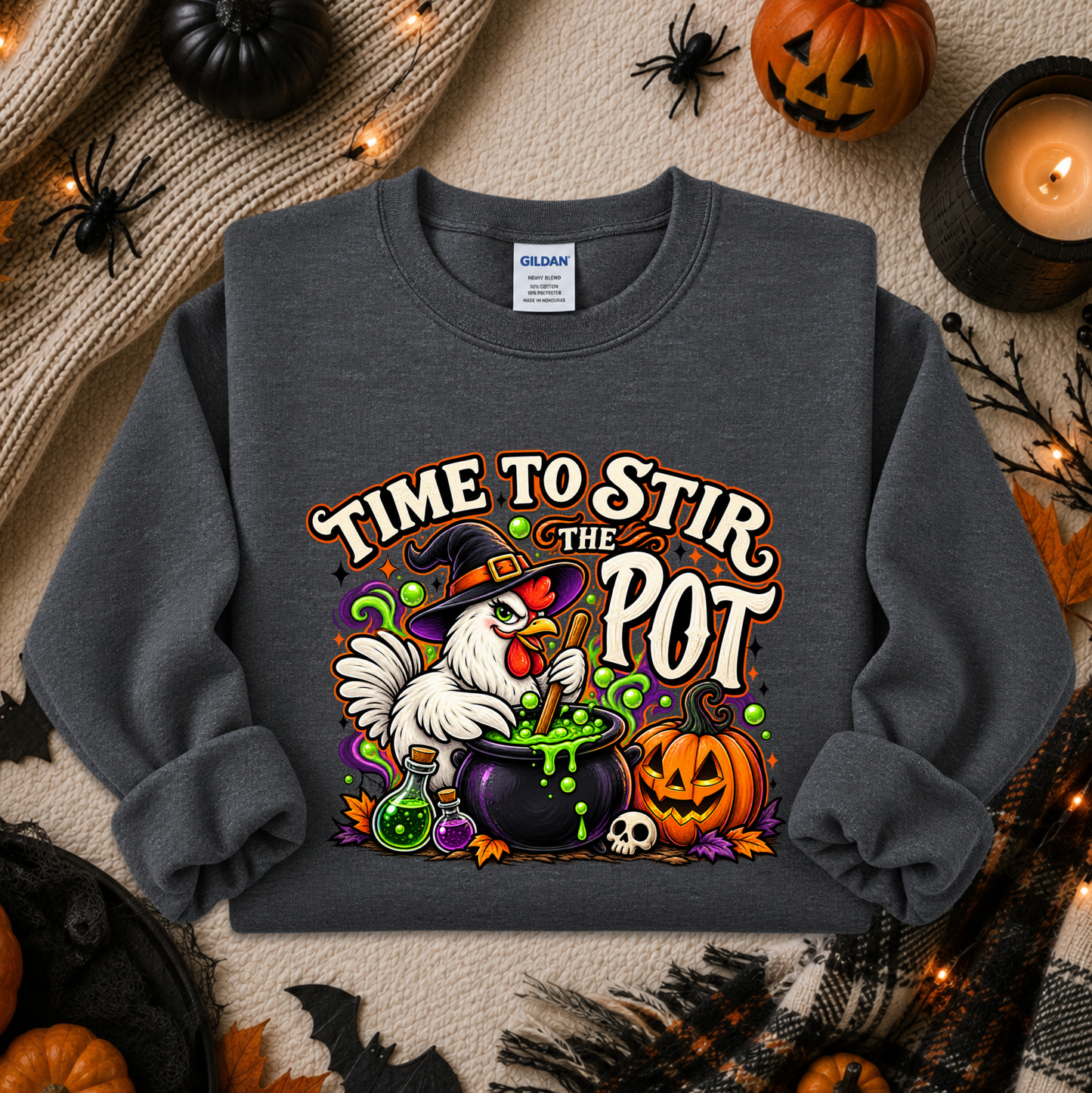 A dark gray crewneck sweatshirt featuring a colorful Halloween graphic of a white chicken stirring a cauldron with the text 'TIME TO STIR THE POT'. The sweatshirt is displayed in a flat-lay arrangement with autumn and Halloween props including pumpkins, a candle, and a knitted blanket.