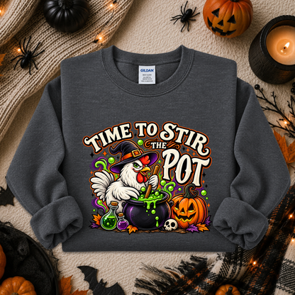 A dark gray crewneck sweatshirt featuring a colorful Halloween graphic of a white chicken stirring a cauldron with the text 'TIME TO STIR THE POT'. The sweatshirt is displayed in a flat-lay arrangement with autumn and Halloween props including pumpkins, a candle, and a knitted blanket.