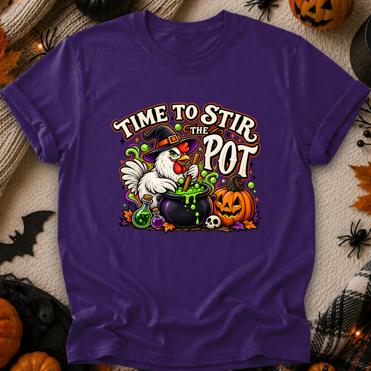 A purple short-sleeve graphic T-shirt laid flat on a textured beige fabric with Halloween decorations around it. The shirt features a colorful front print of a white chicken in a witch hat stirring a cauldron of green potion, with pumpkins, a skull, and the text “TIME TO STIR THE POT.