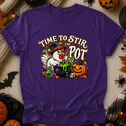 A purple short-sleeve graphic T-shirt laid flat on a textured beige fabric with Halloween decorations around it. The shirt features a colorful front print of a white chicken in a witch hat stirring a cauldron of green potion, with pumpkins, a skull, and the text “TIME TO STIR THE POT.