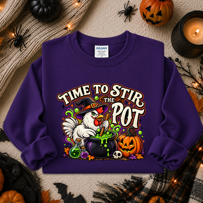 A purple long-sleeve crewneck sweatshirt with a colorful Halloween graphic that reads 'TIME TO STIR THE POT' above a rooster, cauldron, pumpkin, skull, and green slime. The sweatshirt is laid flat on a beige knit blanket surrounded by autumn decorations including pumpkins, a candle, spiderwebs, and fairy lights.