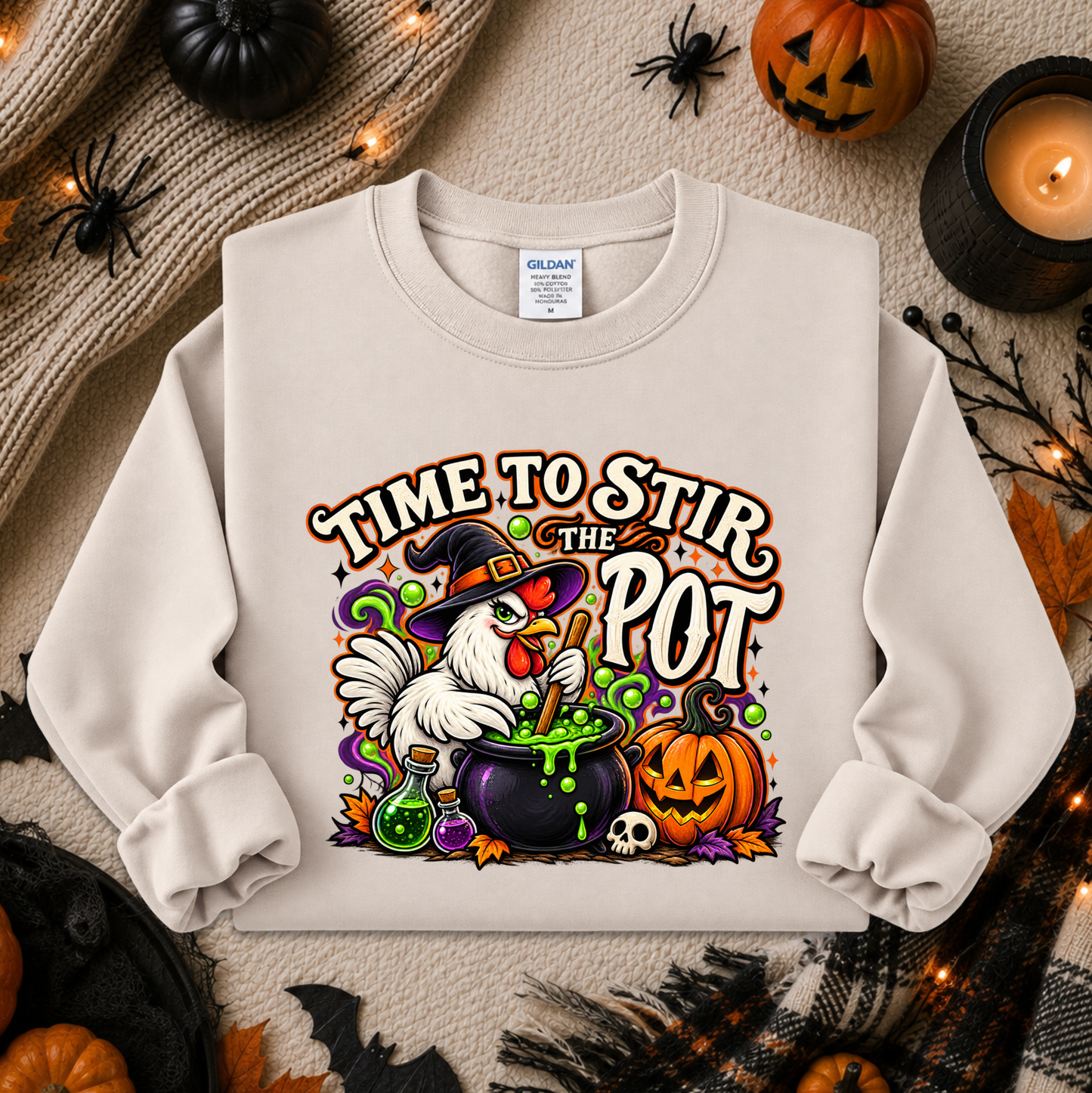 A cream-colored crewneck sweatshirt featuring a colorful Halloween graphic of a white chicken in a witch hat stirring a cauldron with green potion, surrounded by pumpkins, skulls, and the text 'TIME TO STIR THE POT'. The sweatshirt is displayed in a flat-lay arrangement with autumn-themed props including a knitted sweater, candles, and jack-o'-lanterns.