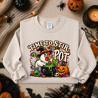 A cream-colored crewneck sweatshirt featuring a colorful Halloween graphic of a white chicken in a witch hat stirring a cauldron with green potion, surrounded by pumpkins, skulls, and the text 'TIME TO STIR THE POT'. The sweatshirt is displayed in a flat-lay arrangement with autumn-themed props including a knitted sweater, candles, and jack-o'-lanterns.