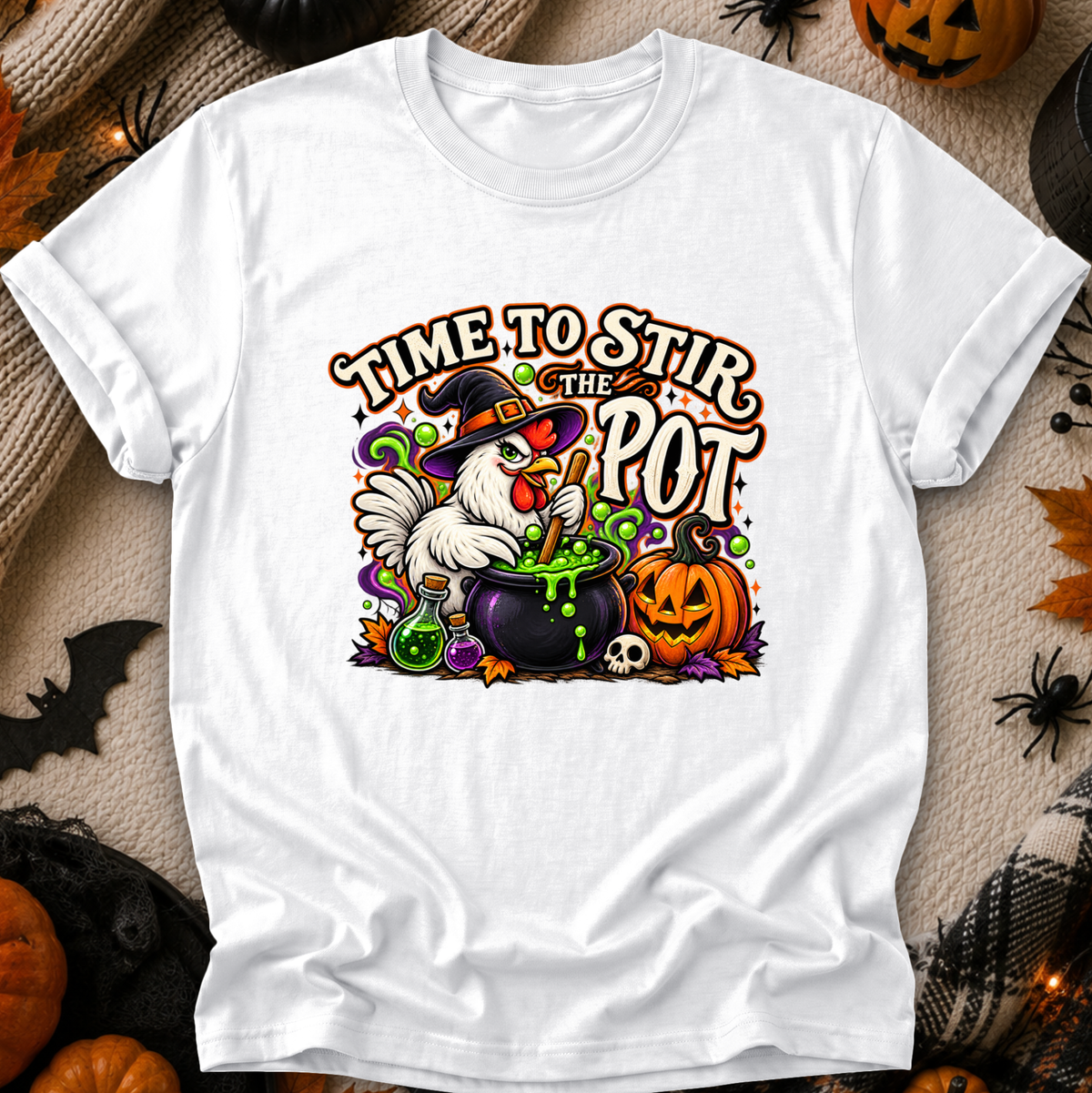 A white short-sleeve graphic T-shirt laid flat on a Halloween-themed background. The shirt features a colorful printed design of a white chicken in a witch hat stirring a cauldron of green potion next to a carved pumpkin, with the text 'TIME TO STIR THE POT.