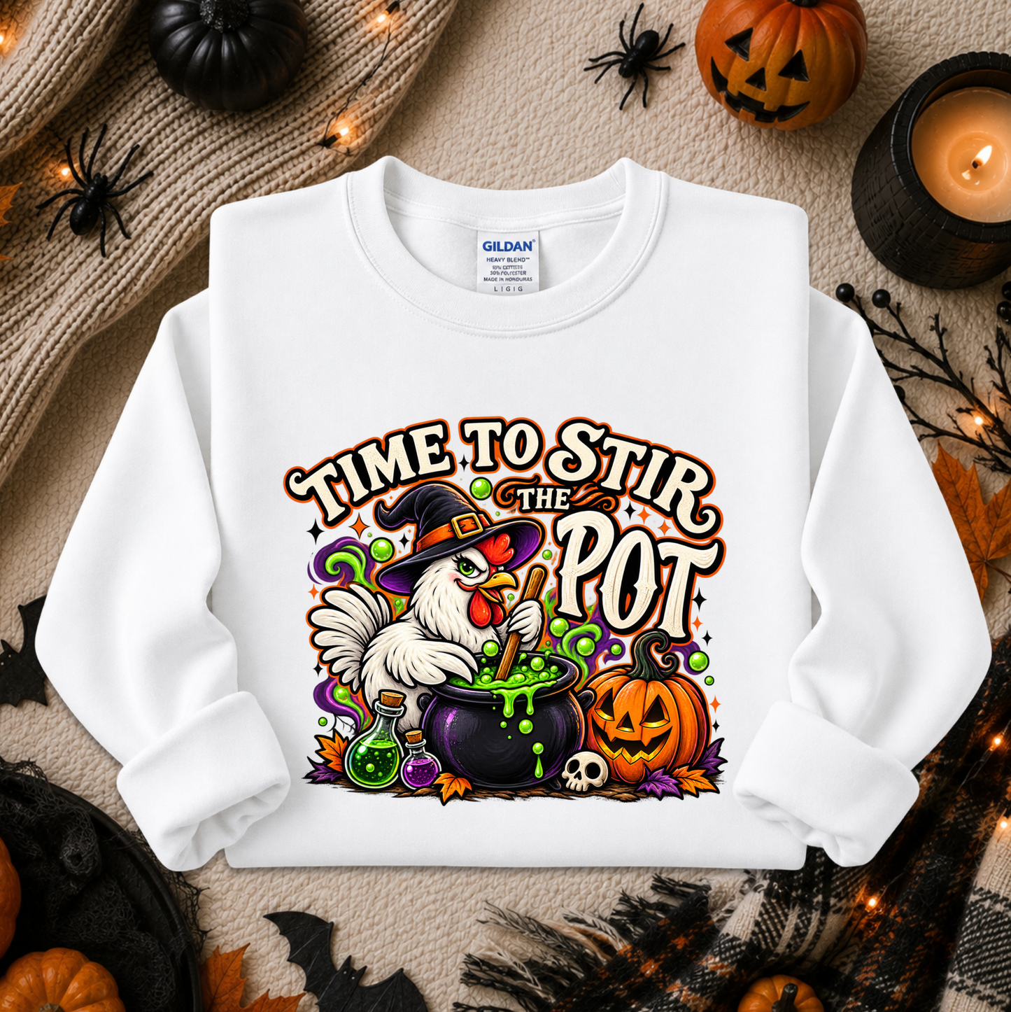 A white long-sleeve crewneck sweatshirt with a colorful Halloween graphic reading 'TIME TO STIR THE POT' and showing a witch chicken, cauldron, pumpkin, skull, and green potion. The sweatshirt is displayed in a flat-lay arrangement with autumn props including pumpkins, a candle, spider decorations, and a cozy knit blanket.