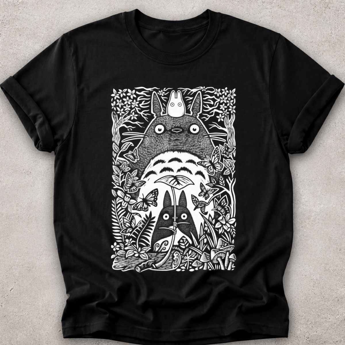 A black short-sleeve crewneck t-shirt featuring a detailed black and white graphic print of Totoro and other My Neighbor Totoro characters surrounded by foliage, laid flat on a textured light gray background.