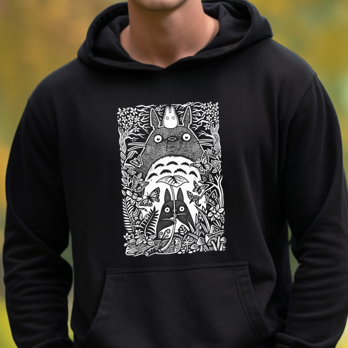 A person wearing a black pullover hoodie with a large white graphic print of Totoro and other My Neighbor Totoro characters on the chest, set against a blurred yellow-green outdoor background.