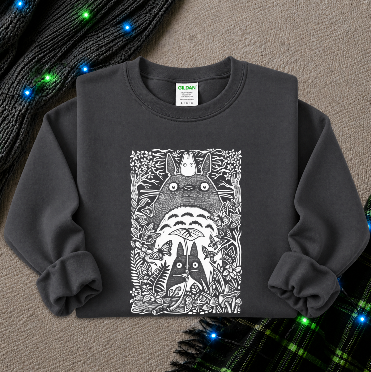 A dark charcoal gray long-sleeve sweatshirt featuring a detailed black and white graphic print of Totoro and other Totoro characters surrounded by foliage. The sweatshirt is laid flat on a textured beige surface with decorative holiday lights and a plaid scarf in the background.