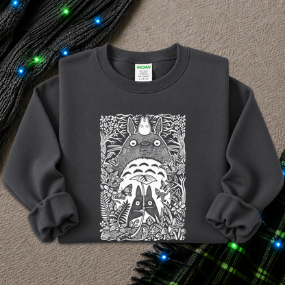 A dark charcoal gray long-sleeve sweatshirt featuring a detailed black and white graphic print of Totoro and other Totoro characters surrounded by foliage. The sweatshirt is laid flat on a textured beige surface with decorative holiday lights and a plaid scarf in the background.