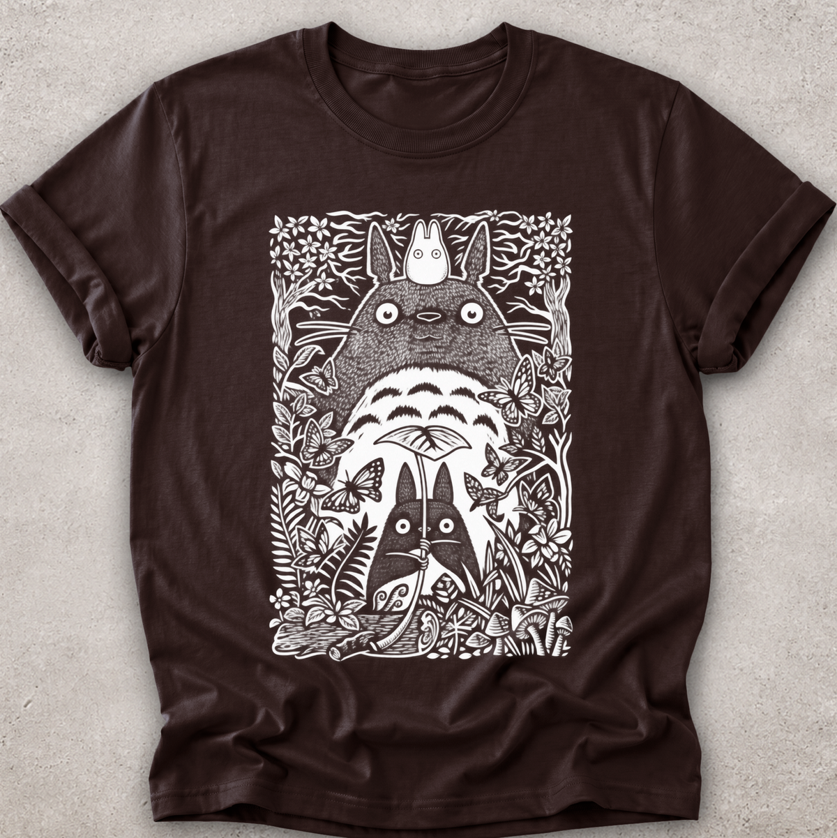 A dark brown short-sleeve t-shirt with a detailed white graphic print of Totoro and other My Neighbor Totoro characters surrounded by foliage, laid flat on a textured gray background.