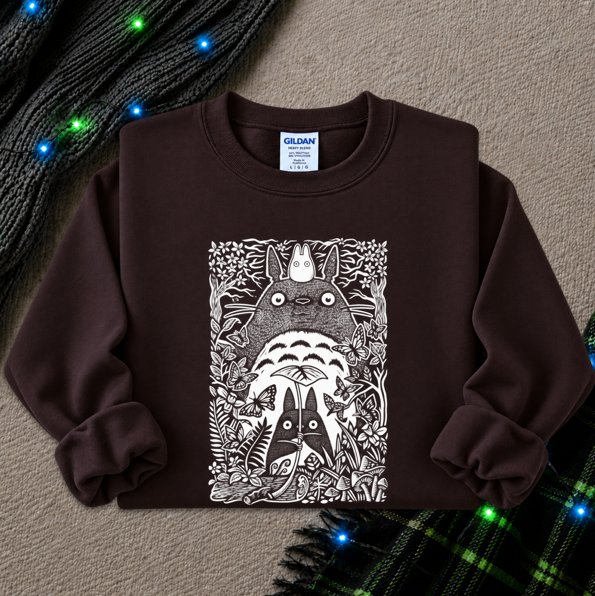 A dark brown crewneck sweatshirt featuring a large white graphic print of Totoro and other My Neighbor Totoro characters, styled flat on a textured beige background with decorative knit items and blue fairy lights.